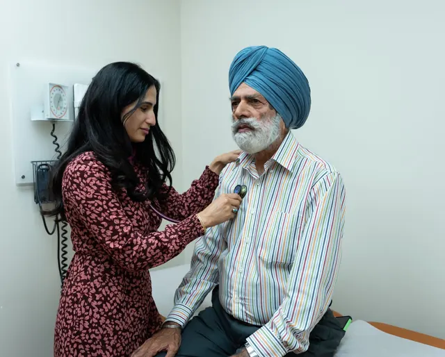 Female doctor listening with a stethoscope to an older Sikh man in a turban seated on an exam couch