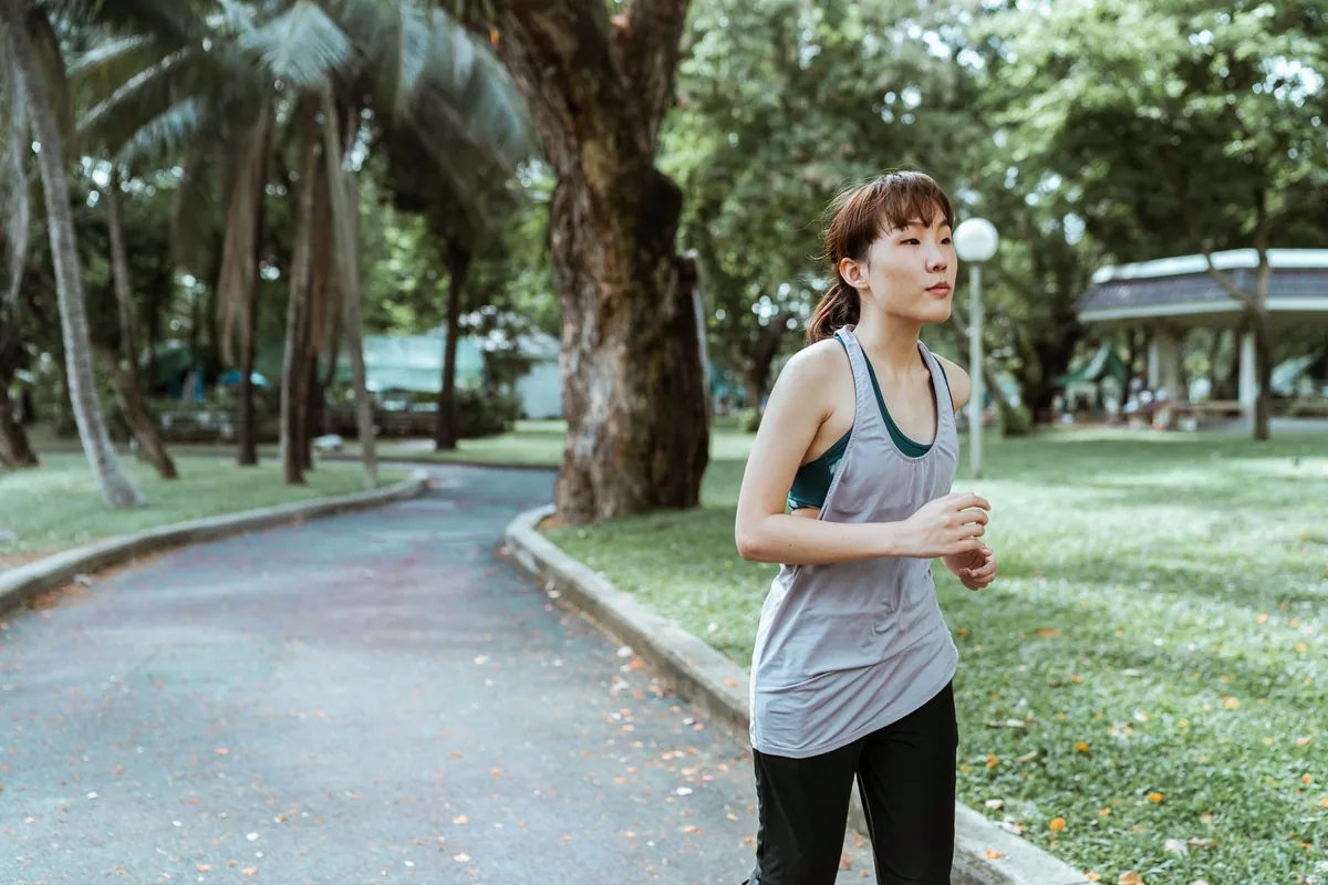 Young Asian woman in a grey vest top jogging along a path in a leafy tropical park
