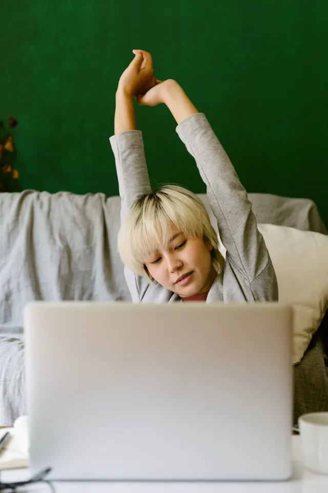 Asian woman with short blonde hair stretching her arms overhead behind a laptop at home