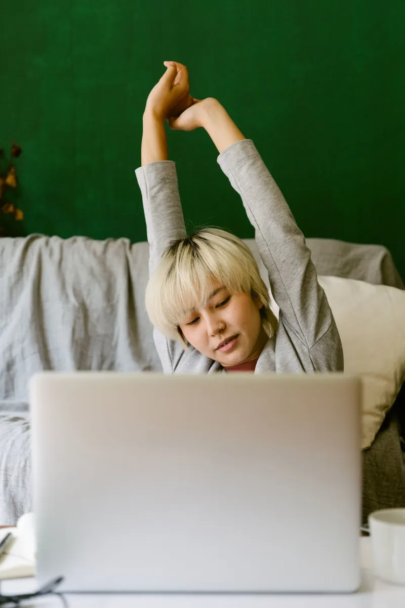 Asian woman with short blonde hair stretching her arms overhead behind a laptop at home
