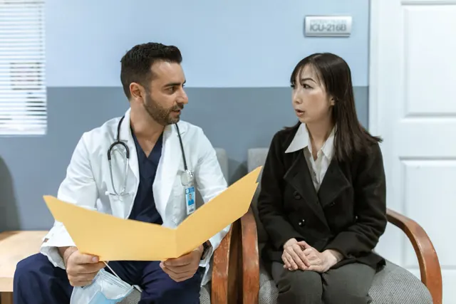 Doctor in a white coat holding an open folder while talking with an attentive Asian woman seated beside him