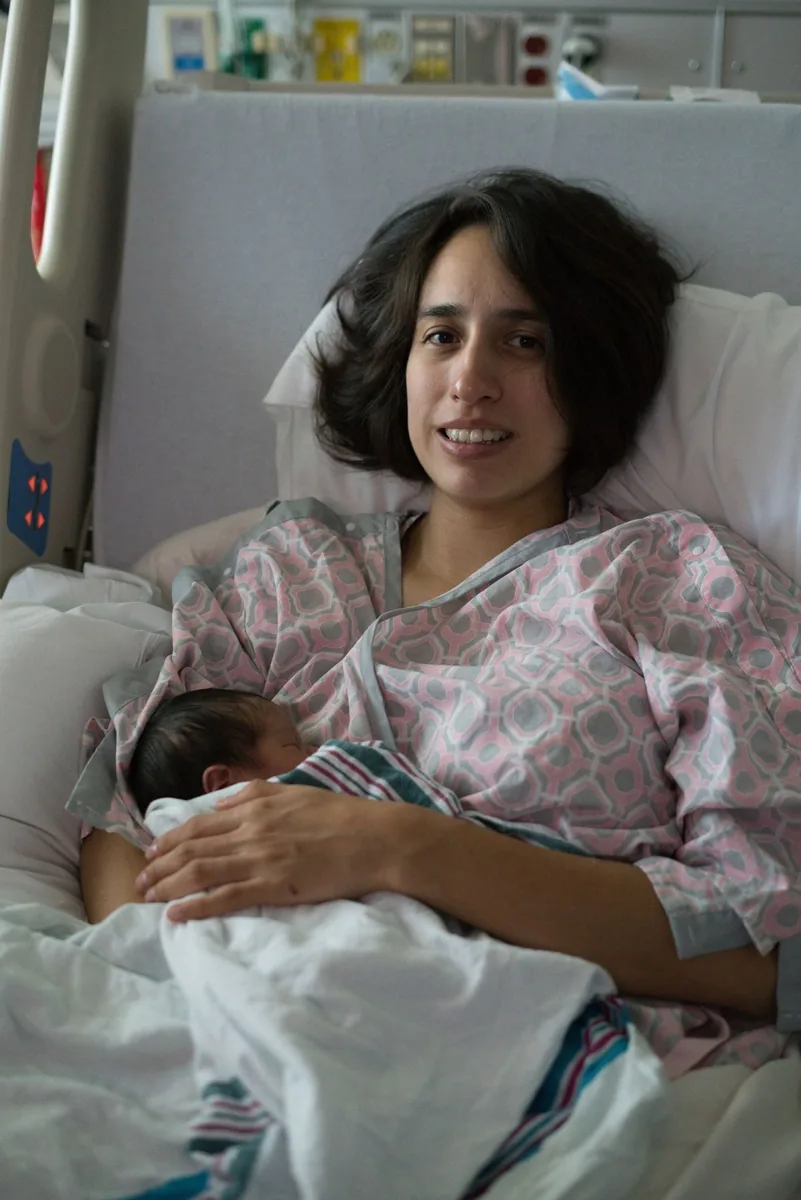 New mother in a hospital gown resting in a hospital bed with her sleeping newborn on her chest