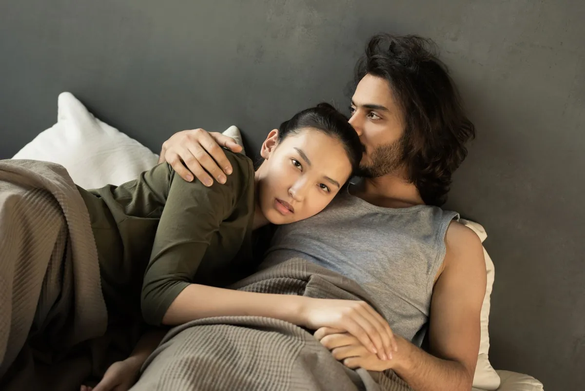 Pasangan berpakaian lengkap beristirahat di tempat tidur, perempuan Asia tampak termenung sementara pasangannya mencium kepalanya