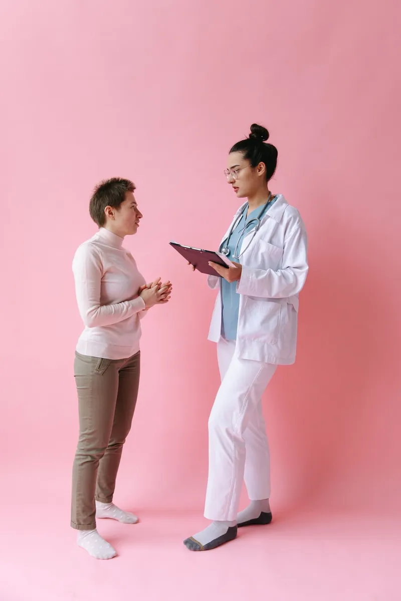 Woman with short hair talking with a female doctor holding a clipboard against a soft pink backdrop