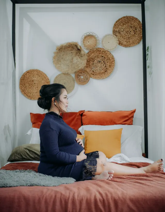 Pregnant Asian woman in a navy knit dress sitting on a bed, both hands resting on her bump, calm expression