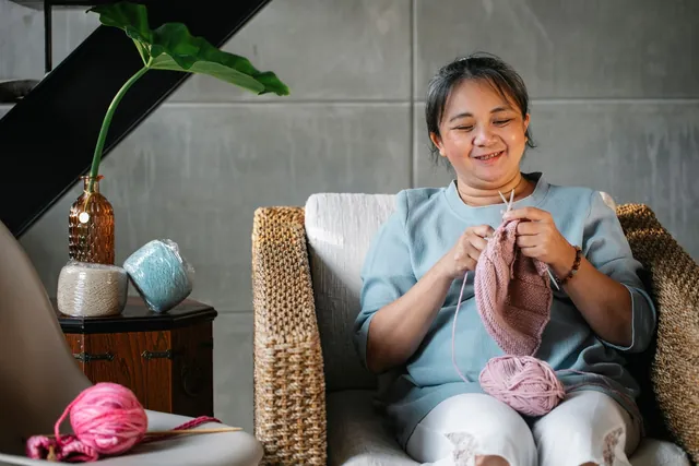 Smiling middle-aged Asian woman knitting with pink yarn in a rattan armchair at home