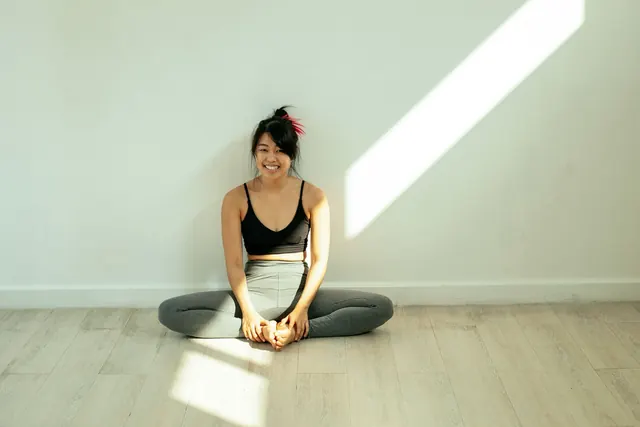 Smiling Asian woman in activewear sitting in a butterfly stretch on a wooden floor in soft sunlight