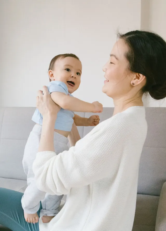 Seorang ibu Asia tersenyum sambil mengangkat bayinya di rumah dalam cahaya siang yang lembut