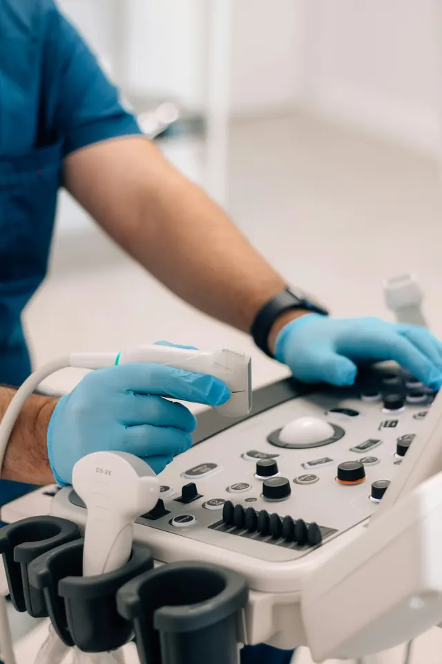 Sonographer in blue gloves holding an ultrasound probe over the control panel of an ultrasound machine
