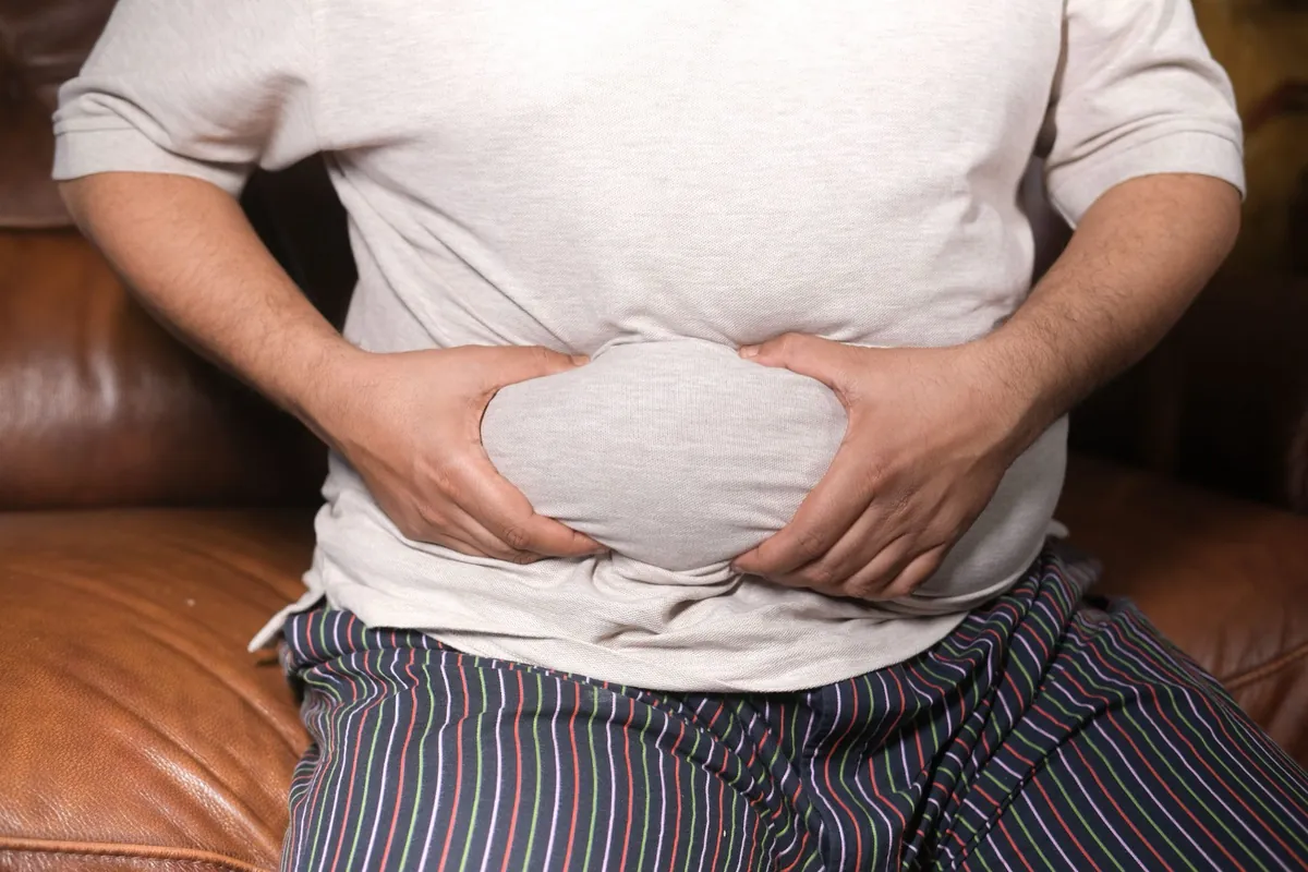Man in a grey T-shirt sitting on a leather sofa with both hands pressed to his lower abdomen, face out of frame