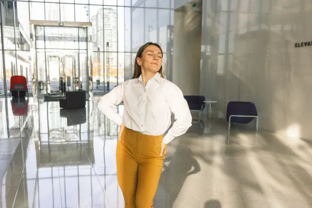Seorang perempuan berpakaian kantor berdiri di lobi yang terang dengan kedua tangan menekan punggung bawahnya, mata terpejam