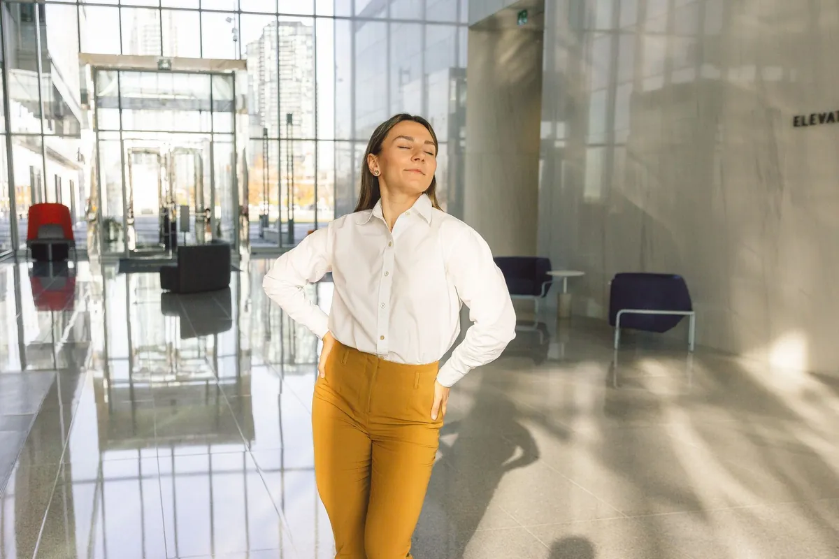 Seorang perempuan berpakaian kantor berdiri di lobi yang terang dengan kedua tangan menekan punggung bawahnya, mata terpejam