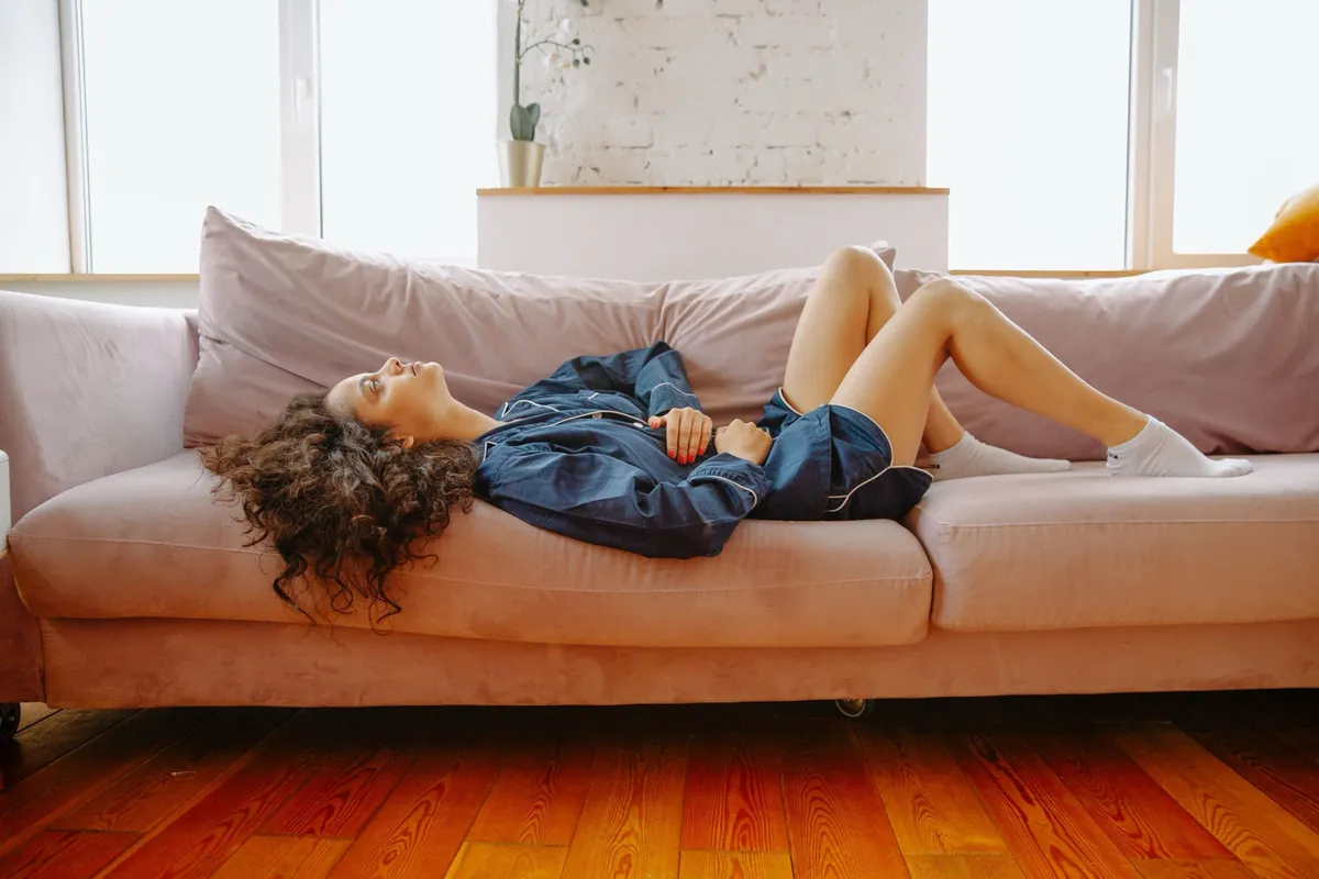 Woman in navy pyjamas lying on a pink sofa with her hands resting on her lower abdomen