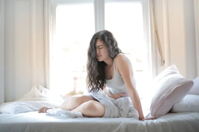 Woman sitting up in bed in bright morning light, one hand on her abdomen, eyes closed in mild discomfort