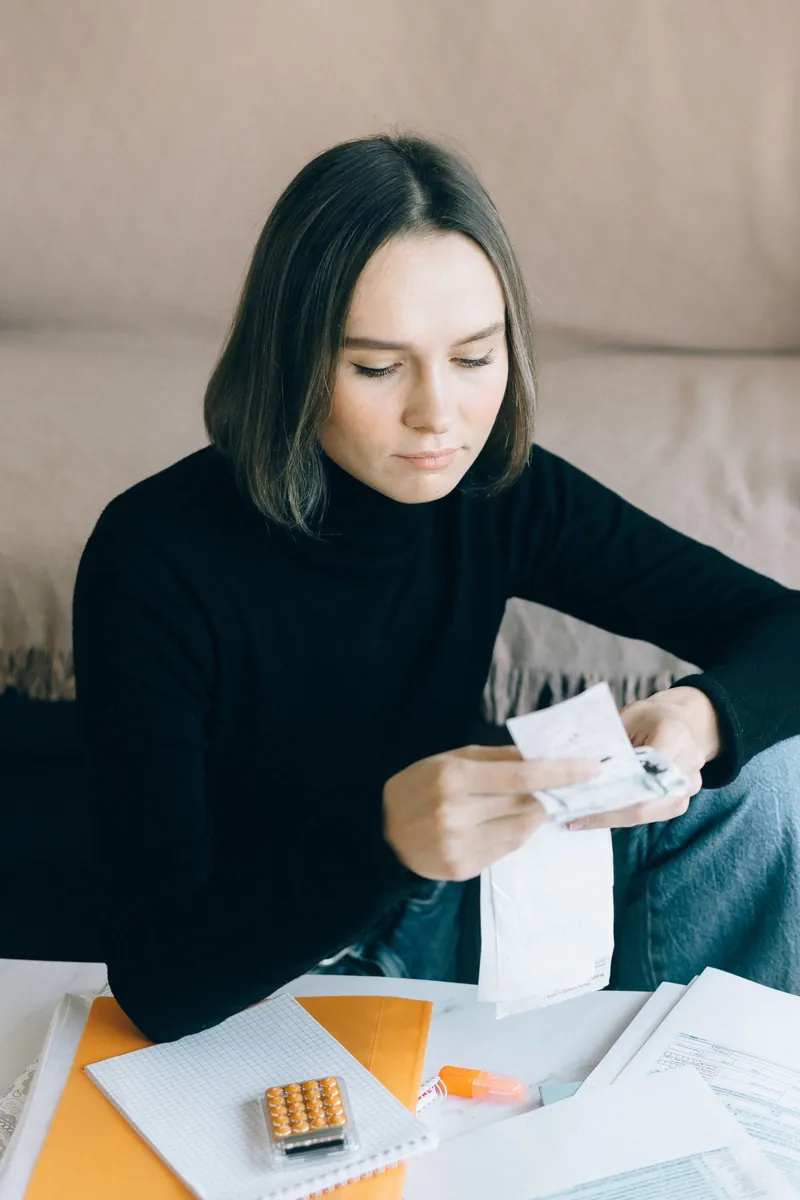 Woman in a black turtleneck reading a long receipt at a table covered with forms and a calculator