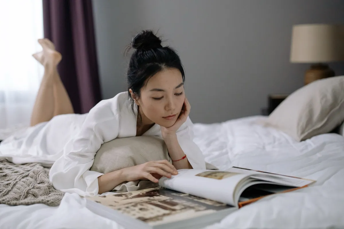 Asian woman in a white robe lying on a bed at home, relaxed and leafing through a large photo book