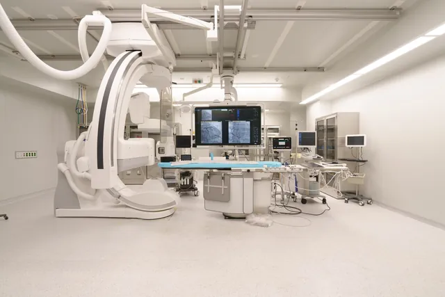 Empty angiography suite with a white C-arm X-ray unit, patient table and overhead screens in a Korean hospital