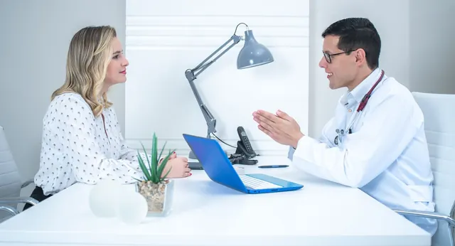 Dokter pria berjas putih menjelaskan sesuatu dengan tangan terbuka kepada seorang perempuan yang tersenyum di seberang meja putih
