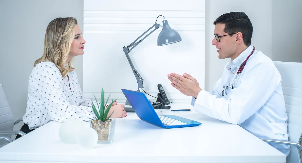 Male doctor in a white coat explaining something with open hands to a smiling woman across a white desk