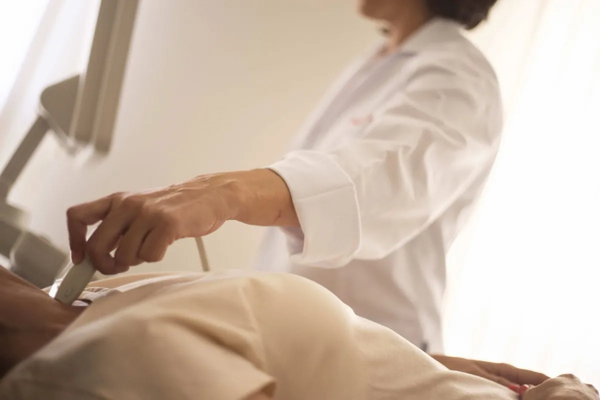 Clinician in a white coat moving an ultrasound probe over the abdomen of a patient lying under a cream gown