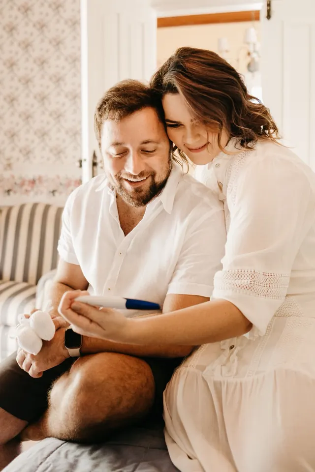 Smiling couple sitting close together at home looking at a pregnancy test, the man holding tiny baby booties