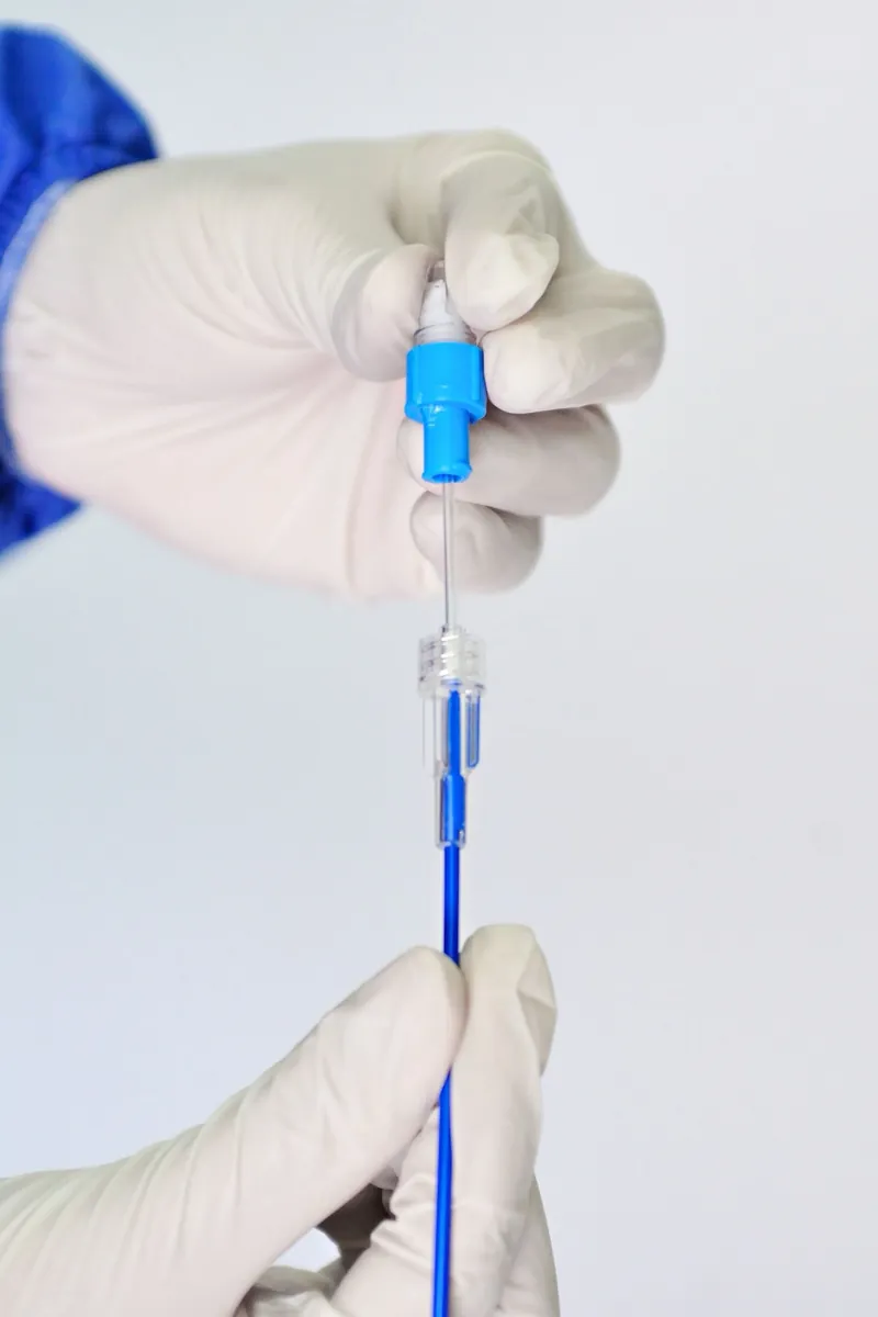 Gloved hands in a sterile blue gown preparing a thin catheter and connector against a plain background