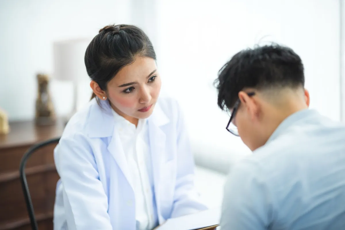 Young Asian man in glasses looking down as a female Asian doctor in a white coat listens with concern