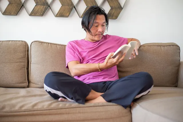 Pria Asia berkaus merah muda duduk bersila di sofa krem di rumah sambil membaca buku