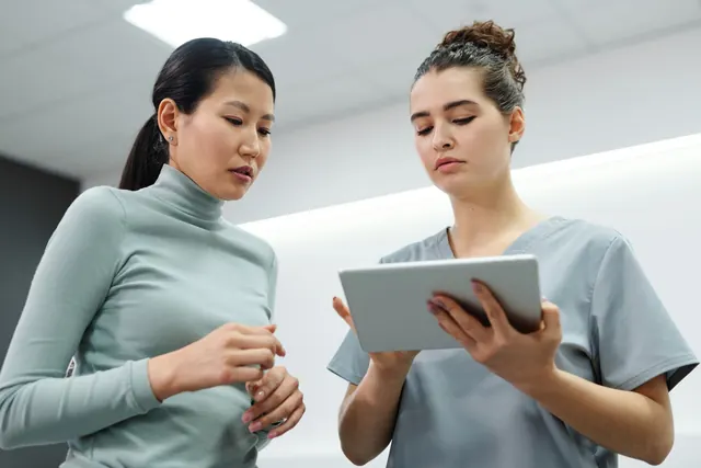 Perempuan Asia dan seorang tenaga kesehatan perempuan berseragam medis melihat tablet bersama di sebuah klinik