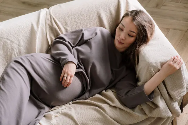 Seorang perempuan Asia yang sedang hamil mengenakan gaun abu-abu longgar beristirahat di sofa dengan tangan di perutnya