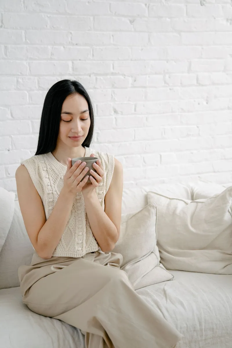 Wanita Asia duduk tenang di sofa putih sambil memegang cangkir hangat dalam cahaya alami yang lembut