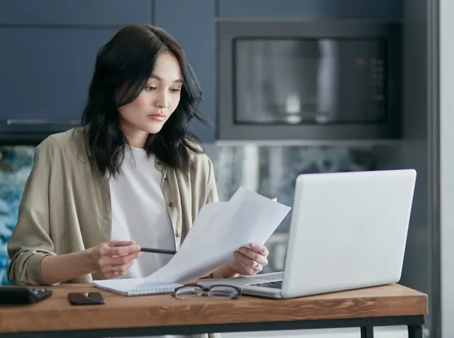 Asian woman at a desk with a laptop, reading through papers with a pen in hand