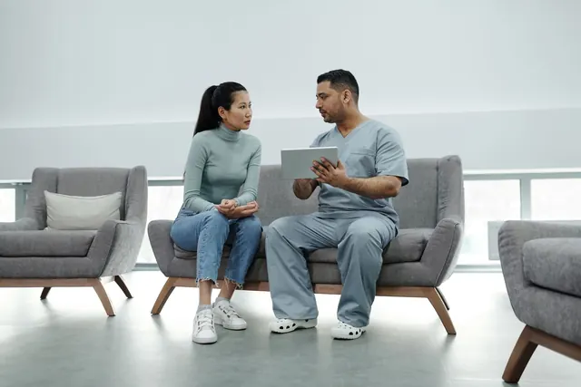 Asian woman and a clinician in scrubs sitting on a sofa in a bright clinic lounge, discussing a tablet