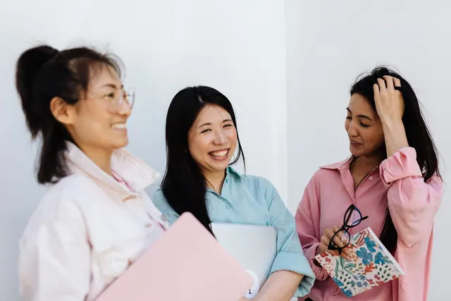 Three smiling Asian women in their 30s and 40s chatting together against a white wall